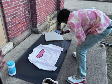 student wearing tye dye pink taking a photo of a white hoodie with pink writing